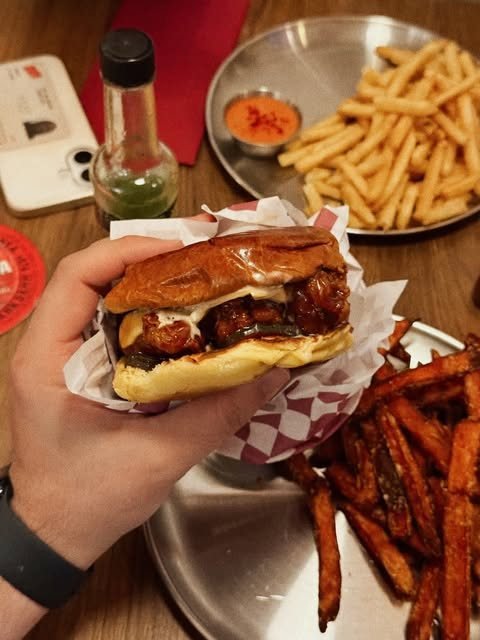 Un bocadillo de pollo frito de POYO sostenido con la mano sobre una bandeja con boniatos fritos y salsas.